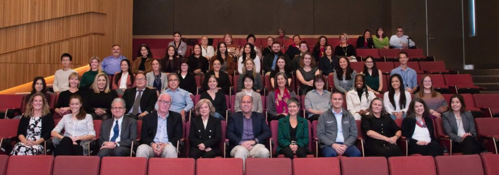 A large group of people seated in an auditorium, arranged in multiple rows, some smiling at the camera.